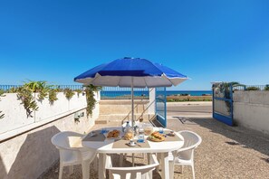 Uma mesa com um guarda-sol azul, cadeiras brancas e vista para o mar.