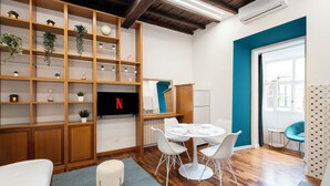 A modern living room with a wooden shelving unit, a dining table with white chairs, a small white sofa, and a TV. - Domus Maggiore Apartment (Roma)