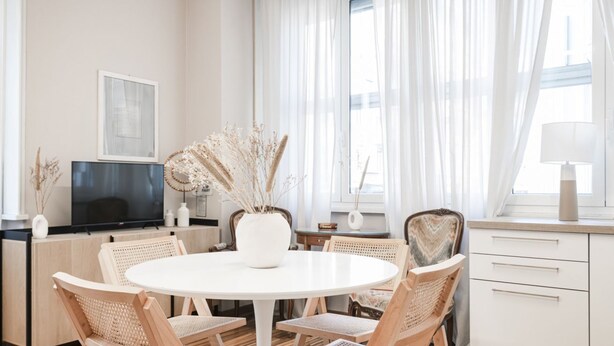 A modern dining area with a round table, wicker chairs, and a white vase with dried plants.