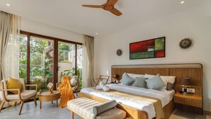 Un dormitorio amplio con una cama grande, cabecera de madera, mesita de noche con lámpara, un banco y una silla. Cuenta con un ventanal con cortinas y vistas al exterior con vegetación.