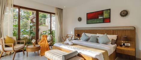 Ein geräumiges Schlafzimmer mit einem großen Bett, einem Holzkopfschild, einem Nachttisch mit Lampe, einer Bank und einem Sessel. Ein großes Fenster mit Vorhängen bietet Blick ins Grüne.