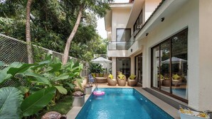 Un área de piscina moderna con una piscina azul cristalina, rodeada de abundante vegetación y mobiliario de exterior.