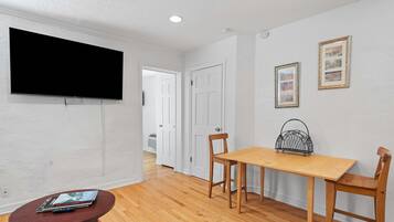A room with a flat-screen TV mounted on the wall, a wooden dining table with two chairs, and a small round table with magazines.