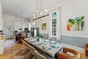 Une cuisine moderne avec des meubles blancs, un coin repas doté d’une longue table dressée pour six personnes, et une grande fenêtre agrémentée de rideaux.