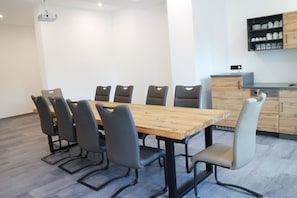 Meetings take place around a spacious table, surrounded by six chairs, in a clean and minimalist room with a projector and kitchen area.