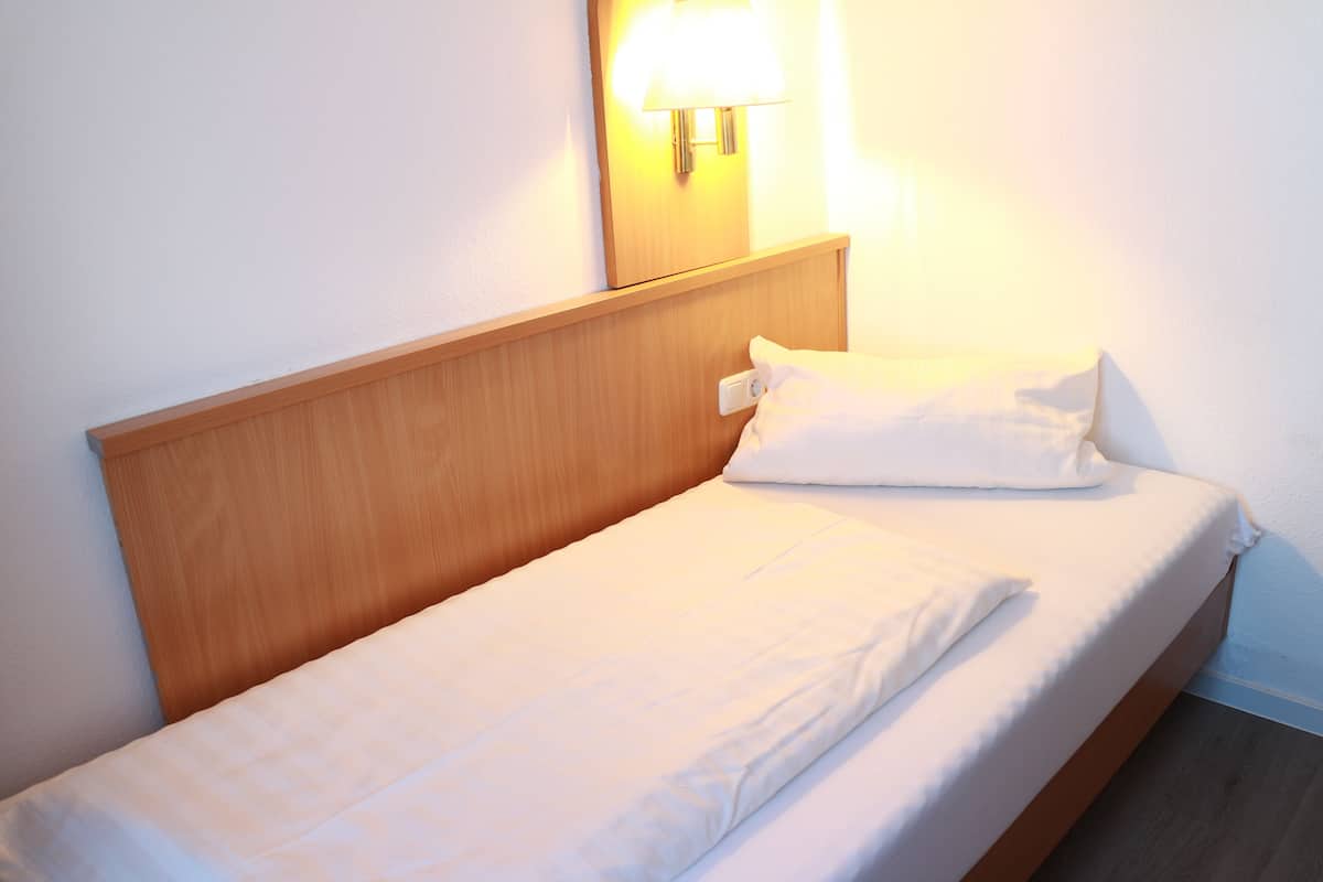 A single bed with a wooden headboard, a white pillow, and a wall-mounted lamp.