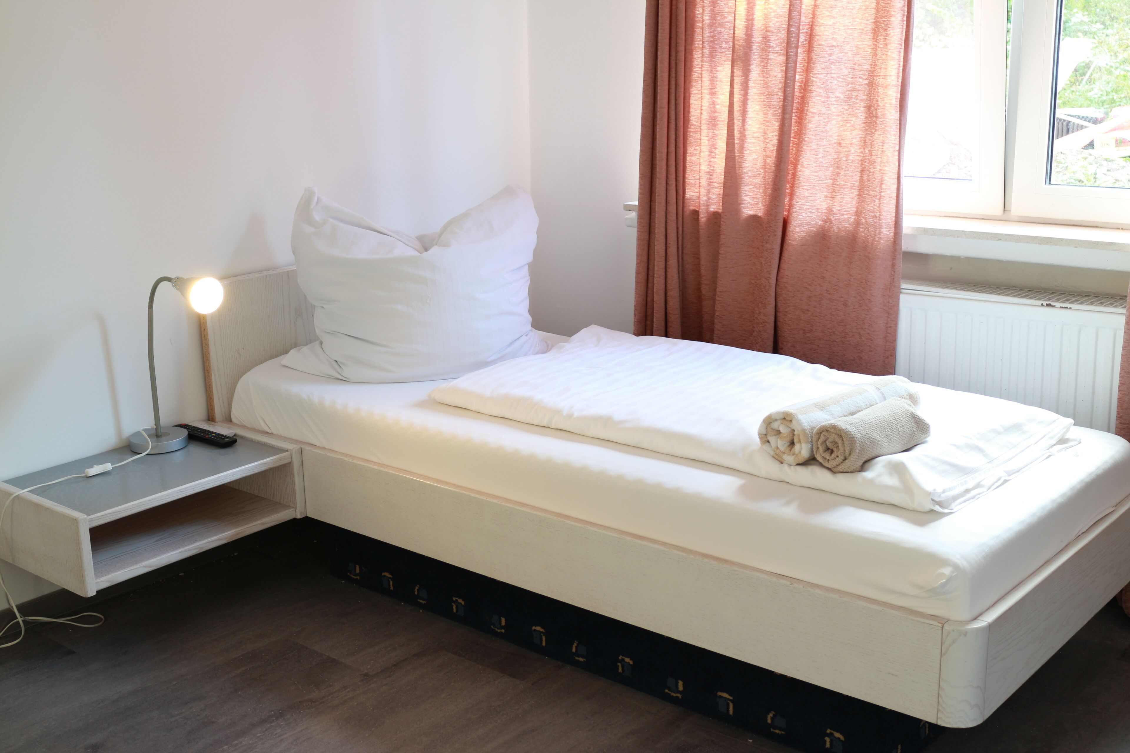 Un lit simple avec un matelas et des oreillers blancs, une table de chevet avec une lampe, et une fenêtre avec des rideaux.