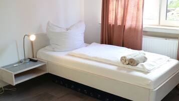 Un lit simple avec un matelas et des oreillers blancs, une table de chevet avec une lampe, et une fenêtre avec des rideaux.