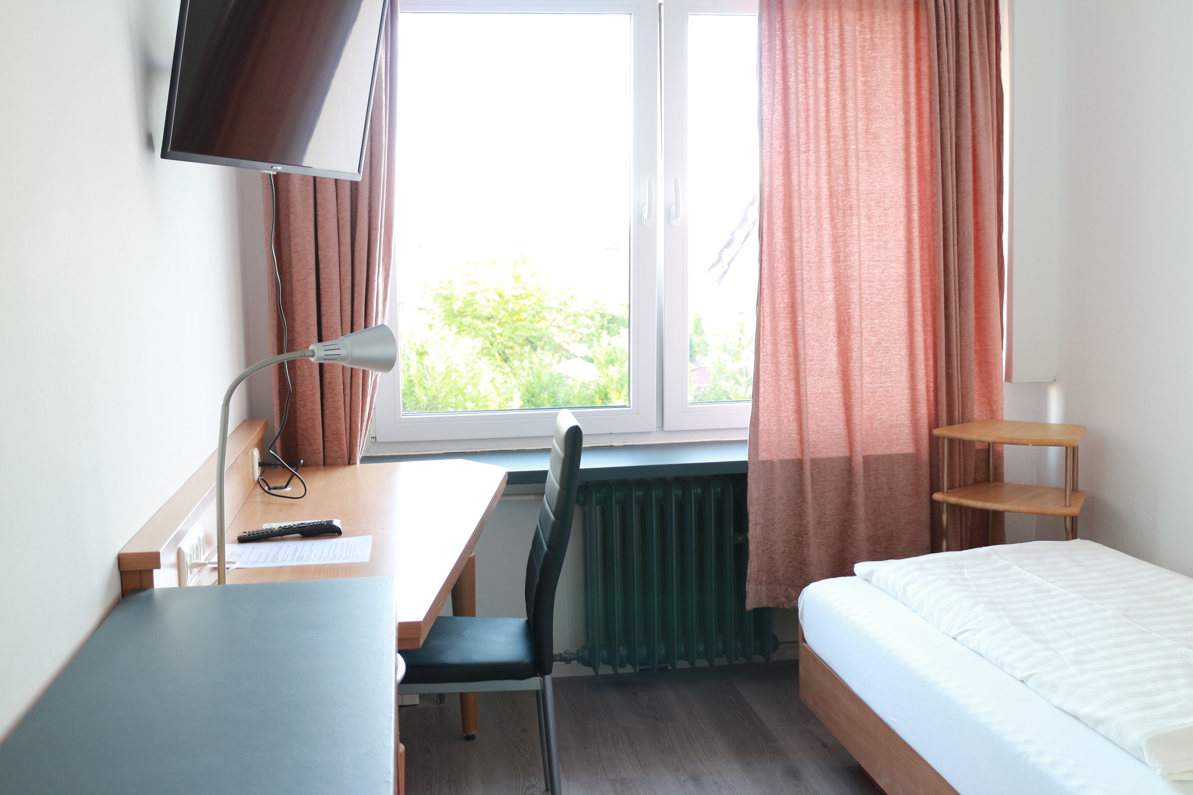 A hotel room with a bed, a desk with a chair, a TV mounted on the wall, and a window with curtains.