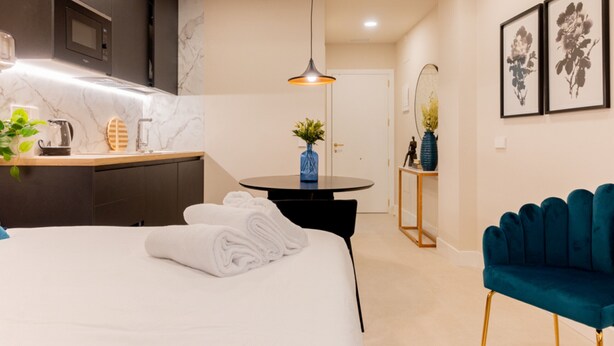 A modern hotel room with a bed, a round table with a vase, a blue chair, a kitchen area with a microwave, and a mirror with a vase. - Estudio Oriente - Design et confort dans DELICIAS MADRID (Madrid)