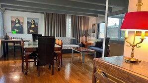 Ein modernes Wohnzimmer mit Holzboden, einer Essecke mit Tisch und Stühlen, einem Sofa, einem Couchtisch und einer Lampe. An der Wand hängen zwei Bilder und ein Fenster mit Vorhängen.