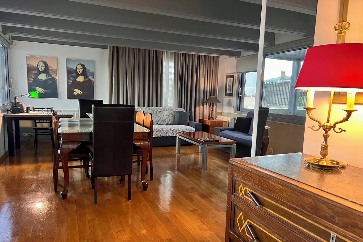 Una sala de estar moderna con suelo de madera, un comedor con mesa y sillas, un sofá, una mesa de centro y una lámpara. Hay dos cuadros en la pared y una ventana con cortinas.