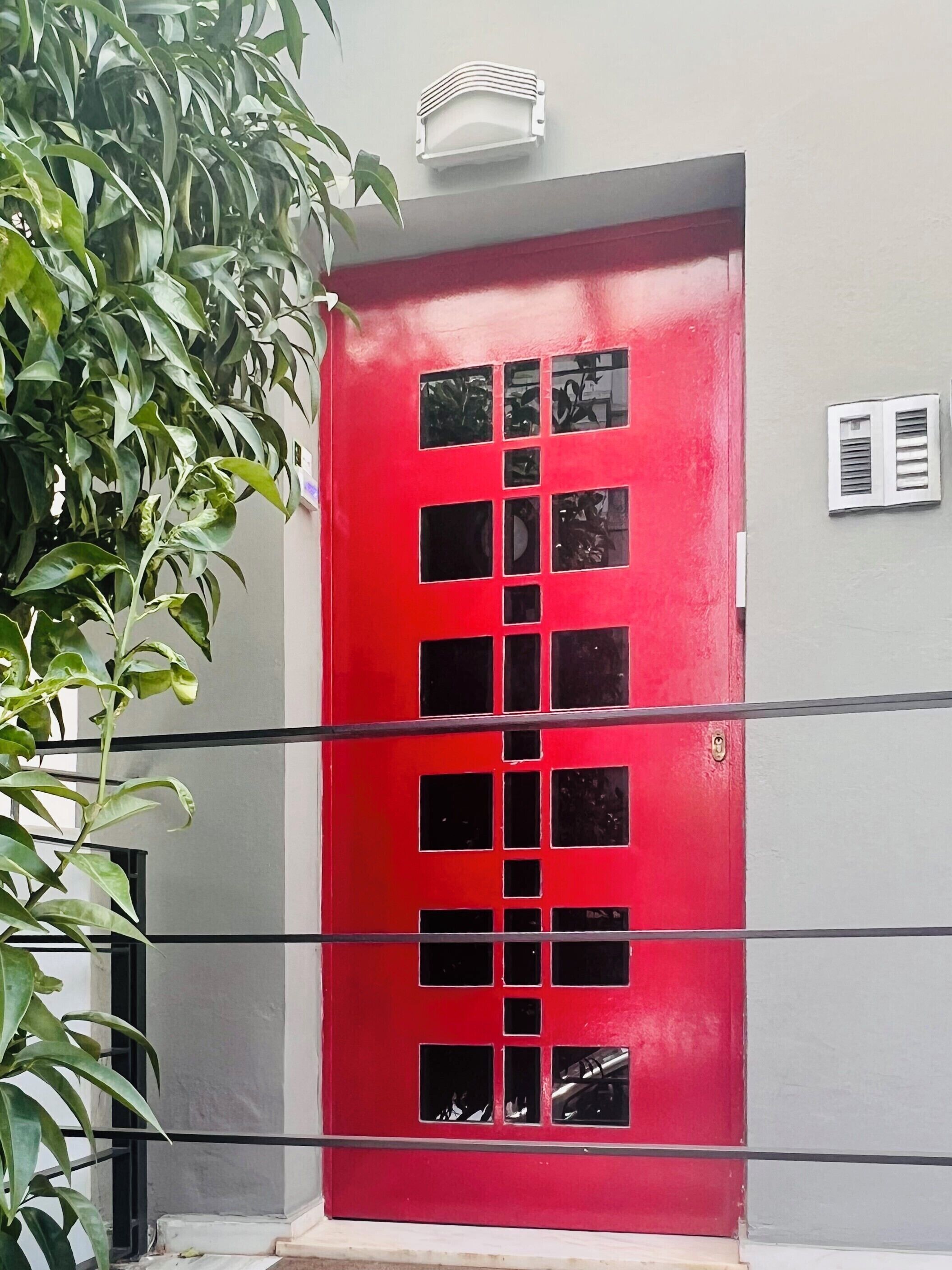 Une porte rouge ornée de plusieurs fenêtres carrées, un mur blanc et un petit appareil de climatisation placé au-dessus.