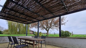An outdoor patio with a wooden table and chairs, a thatched roof, and a view of a tree and grassy field. - Cozy cottage in Athienville (Athienville)
