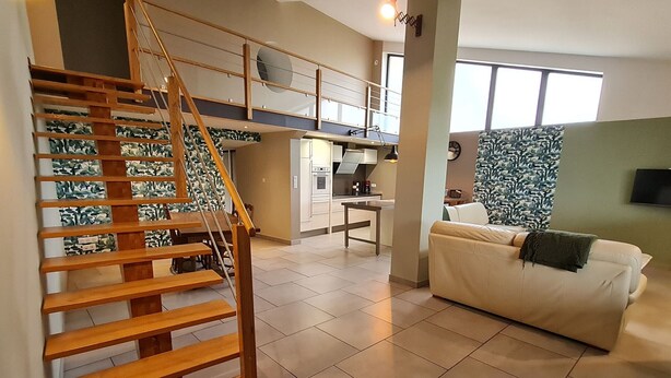 A modern living room with a wooden staircase, a sofa, a kitchen area, and a large window. - Cozy cottage in Athienville (Athienville)