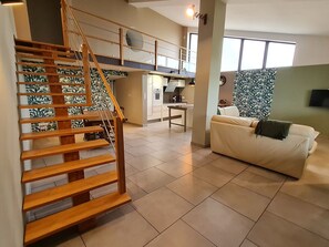 Un salon moderne avec un escalier en bois, un canapé, un coin cuisine et une grande fenêtre.