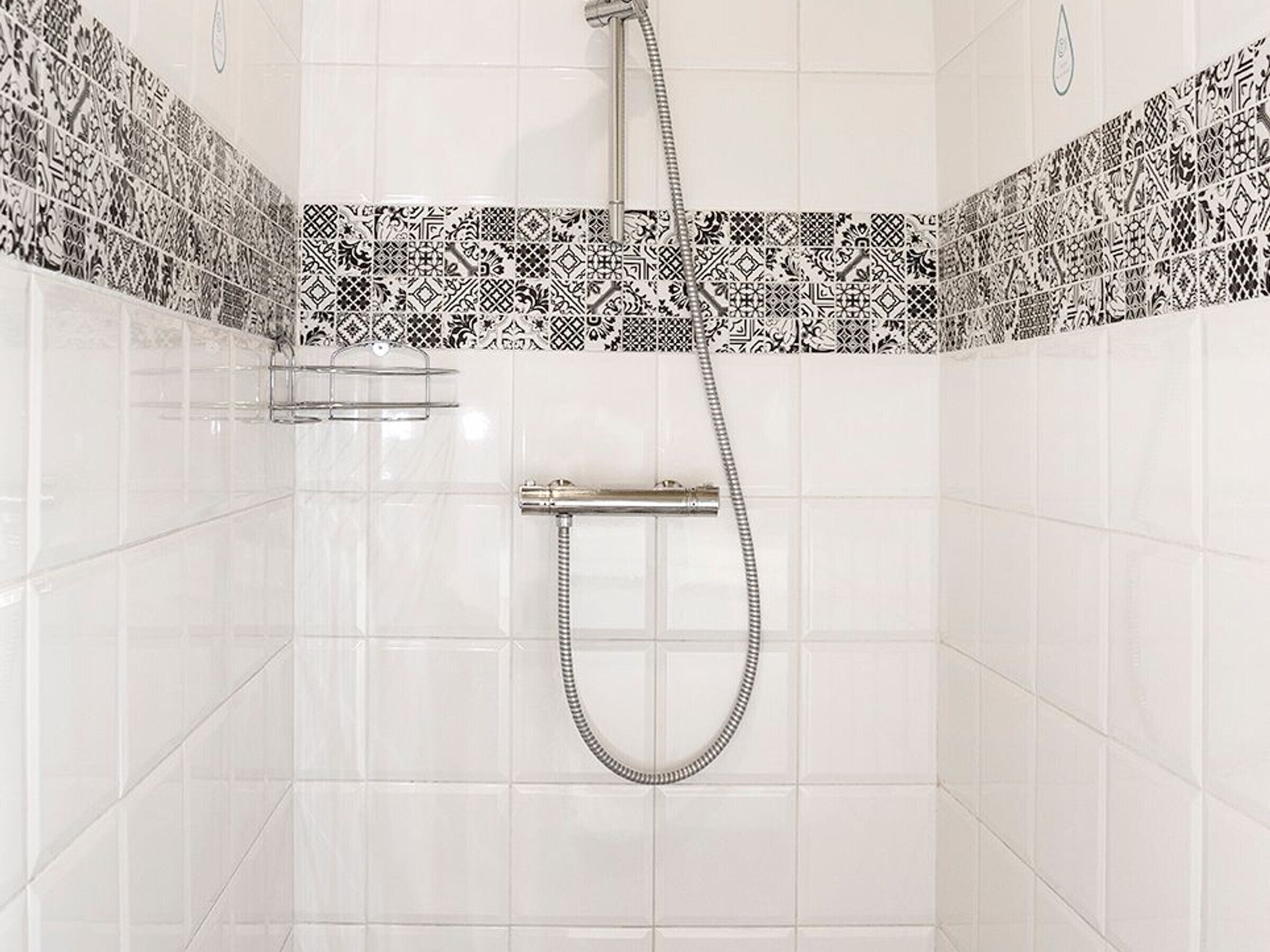 Un baño con azulejos blancos, ducha con manguera y un zócalo de baldosas estampadas.