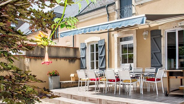Une terrasse avec des sièges extérieurs, un espace abrité et une vue sur un bâtiment doté de fenêtres et de volets.