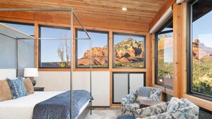 Quarto com cama, poltrona, mesa e amplas janelas com vista para o deserto.