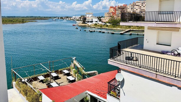 Ein Objekt direkt am Wasser mit einem Deck mit rotem Dach, Außenmöbeln und Blick auf das Wasser und die Gebäude.