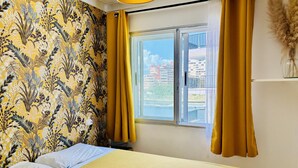Ein Zimmer mit einem Bett, gelben Vorhängen, einem Fenster mit Blick auf Gebäude und einer Wand mit Muster.