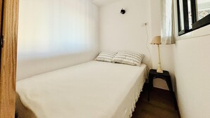 A small, white, single-bed room with a bedside table and lamp, a window with curtains, and a wall socket. - Apartment in Roses, near beach, pet-friendly (Roses)