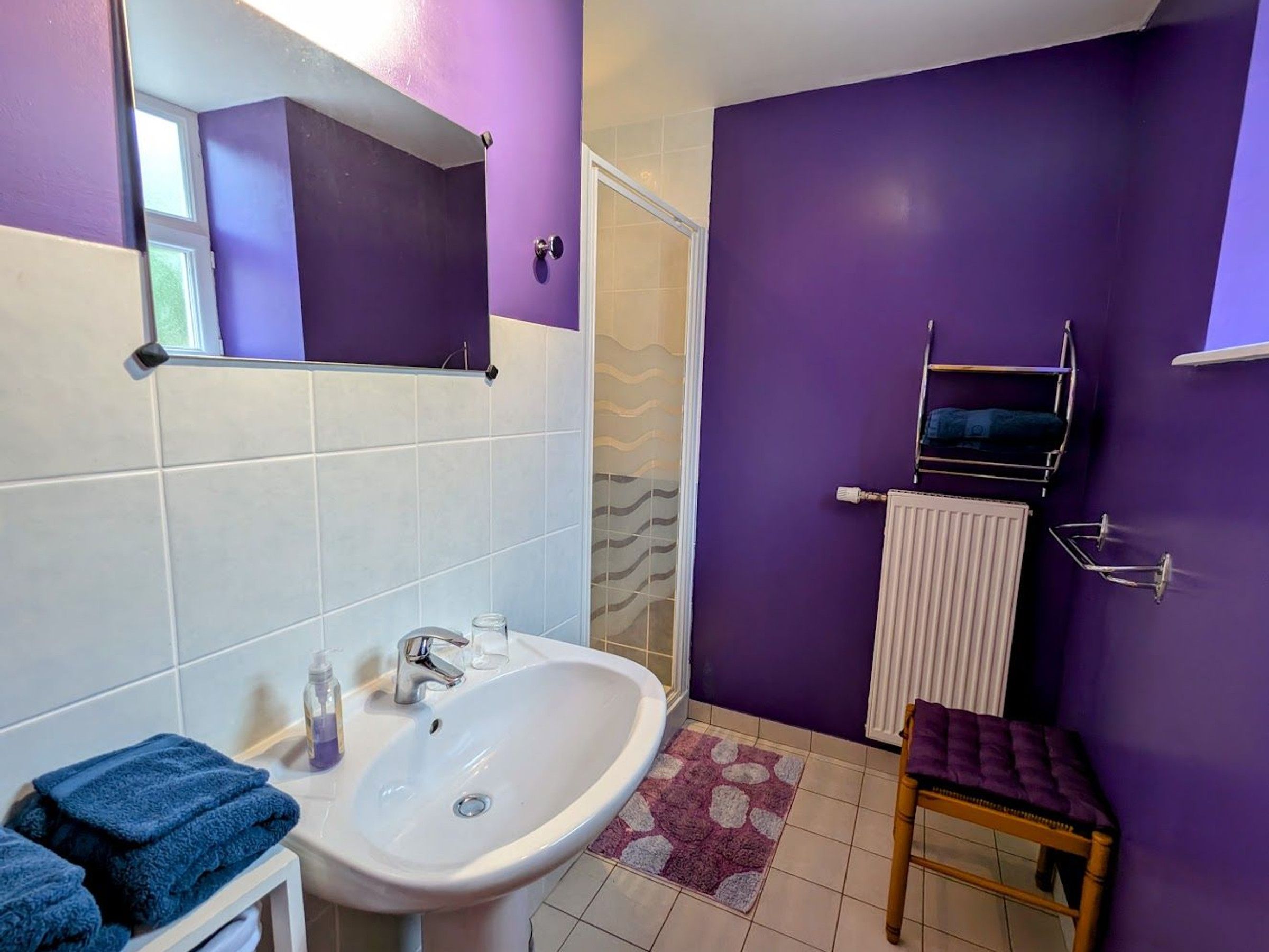 A bathroom with a white sink, a mirror, and a purple wall. There is a towel rack and a small wooden stool.