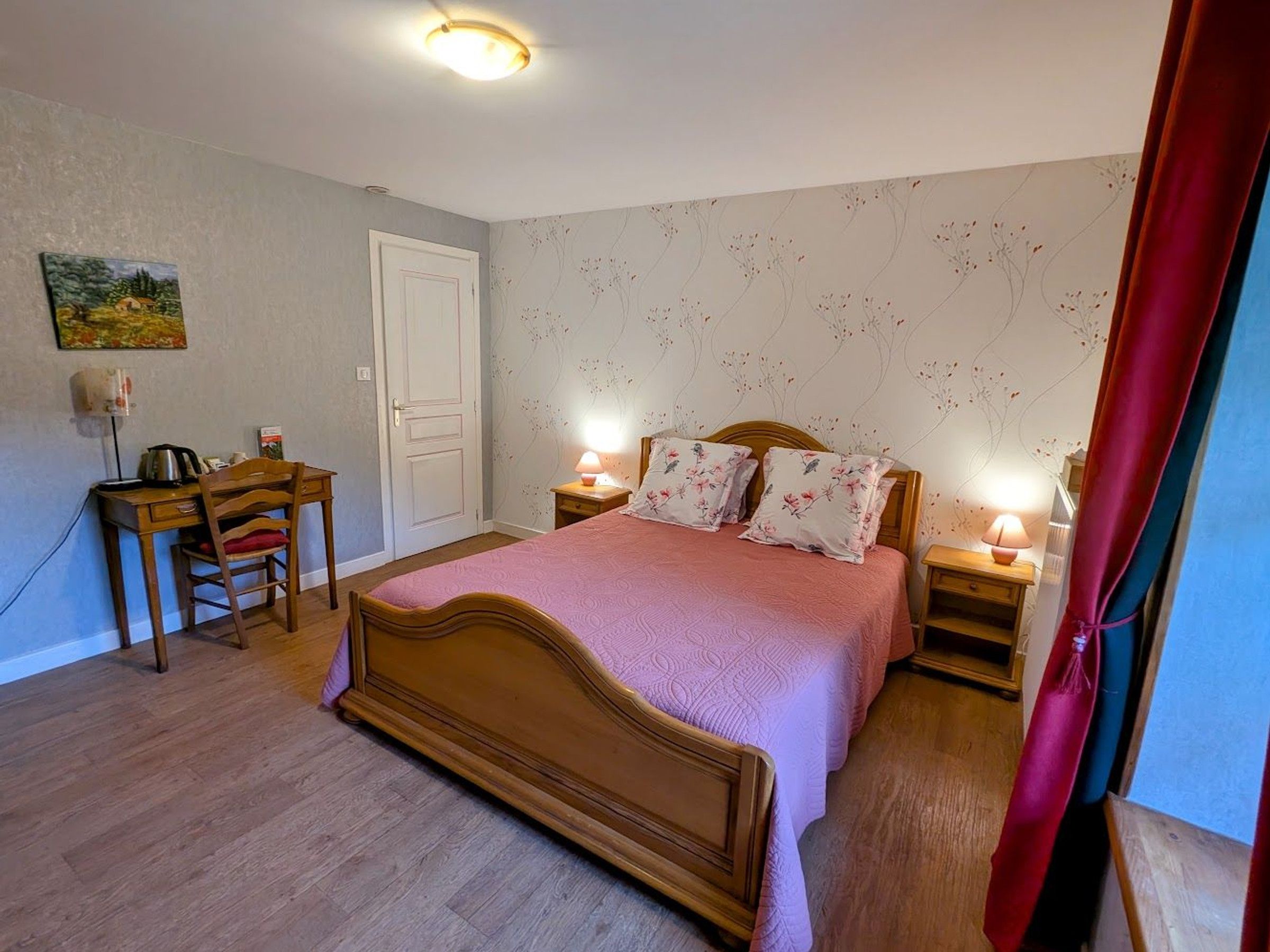 A bedroom with a wooden bed, a desk with a chair, two bedside lamps, and a door.