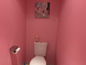 Une salle de bain avec des murs roses, des toilettes blanches, un porte-rouleau de papier toilette et un petit flacon posé sur le réservoir des toilettes.