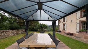 Un espacio de comedor al aire libre cubierto, con una mesa y sillas de madera, un muro de piedra y un edificio al fondo.