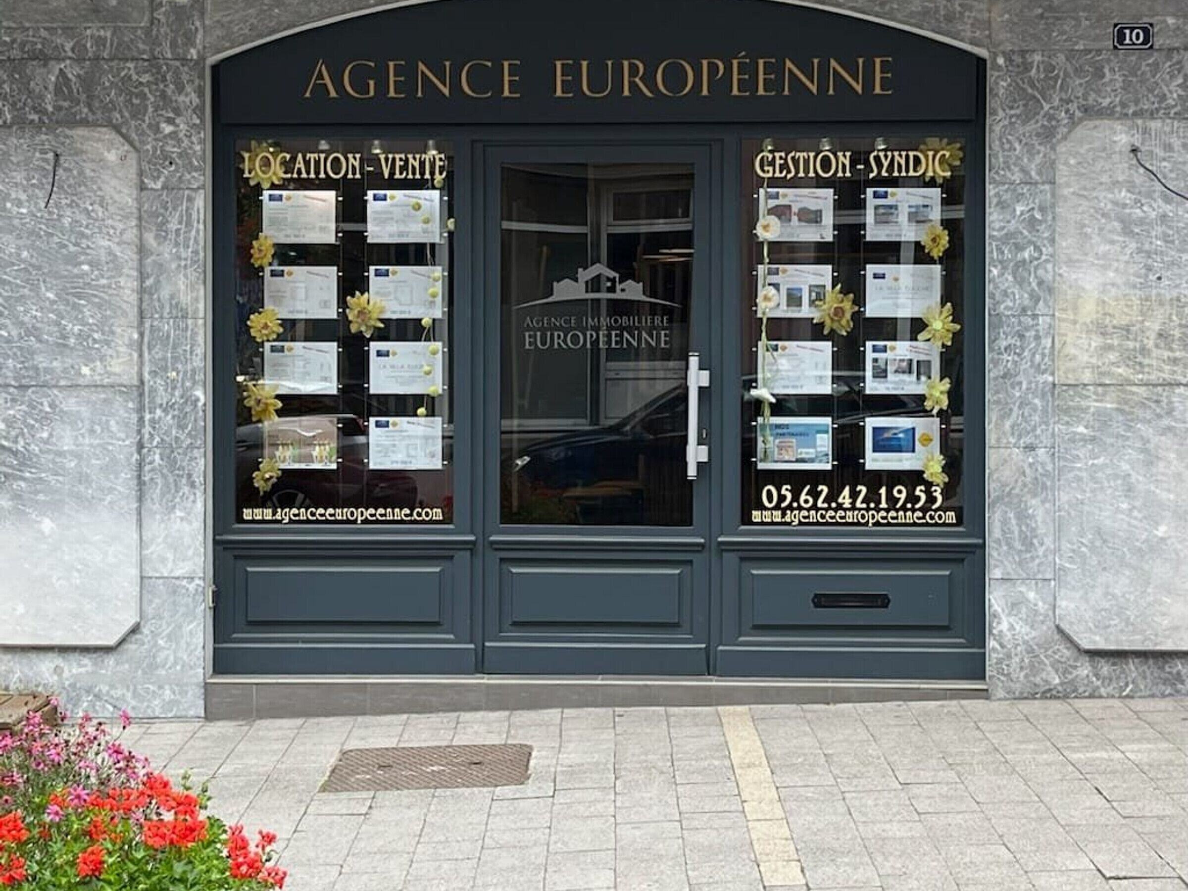 The entrance to AGENCE EUROPEENNE with two glass doors, each adorned with framed documents and yellow flowers.