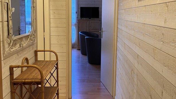 A narrow hallway with wooden paneling, a wicker chair, a television in the background, and a door leading to another room.