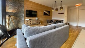 En moderne stue med en stenvæg, træpaneler, en stor grå sofa, et fladskærms-tv og et spiseområde med et træbord og stole.