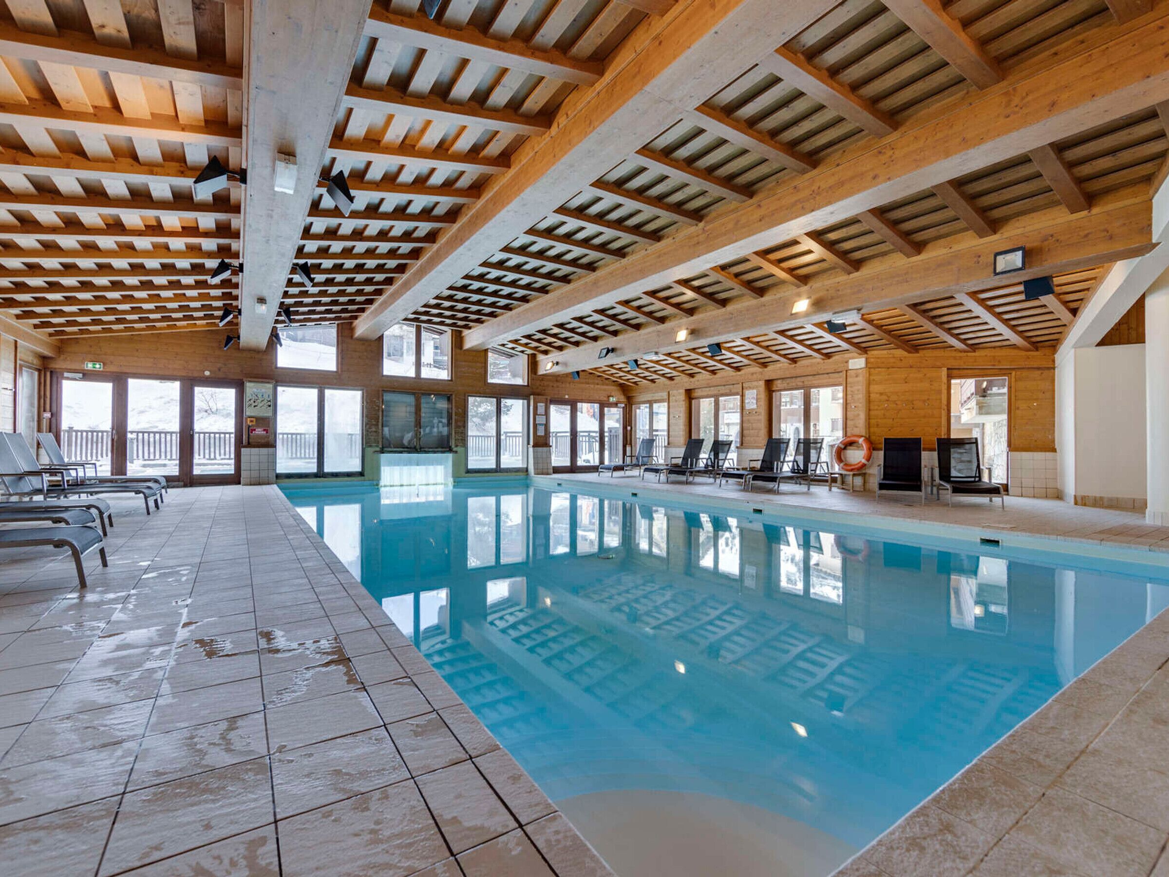 Ein überdachter Pool mit Holzbalken, gefliestem Boden und sonnendurchfluteten Fenstern.