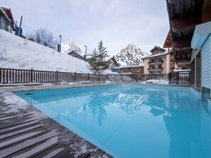 Una piscina blava i cristal·lina envoltada per un pont de fusta, amb muntanyes cobertes de neu i edificis al fons.