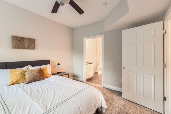 A bedroom with a bed, a ceiling fan, a door, and a hallway leading to a bathroom.