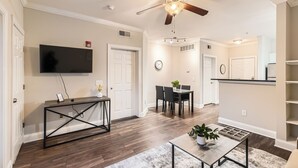 A modern living room with a ceiling fan, a flat-screen TV, a dining area with a table and chairs, and a kitchen area with cabinets and a countertop.