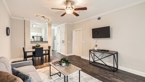 A modern living room with a ceiling fan, a flat-screen TV, a dining area with a kitchen in the background, and a cozy seating area with a coffee table.