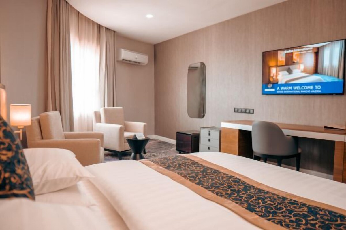 A hotel room with a bed, two armchairs, a desk, and a television displaying a welcome message.