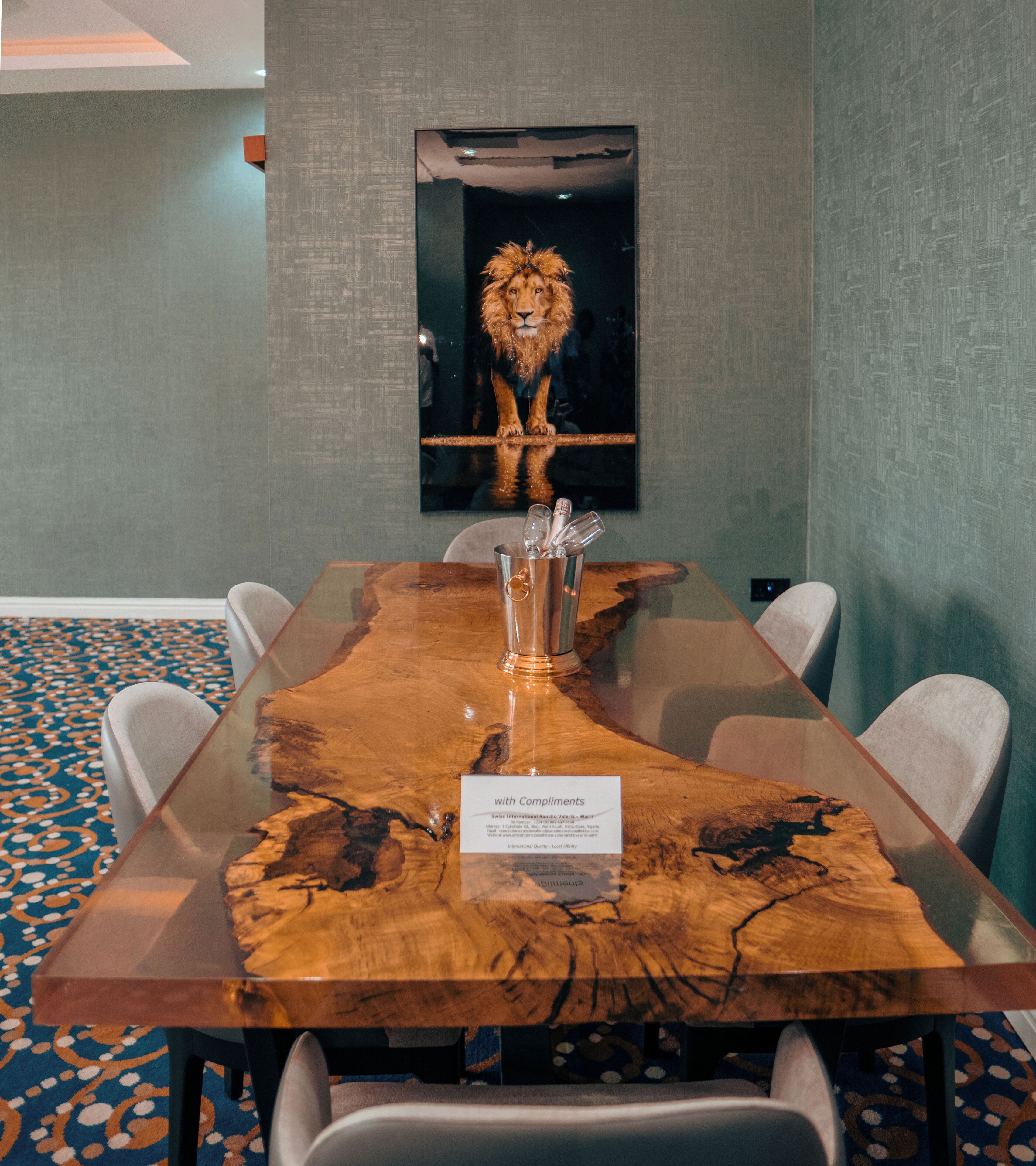 Una mesa de conferencias con una superficie de madera singular, rodeada de sillas, en una sala con alfombra estampada y un cuadro en la pared.