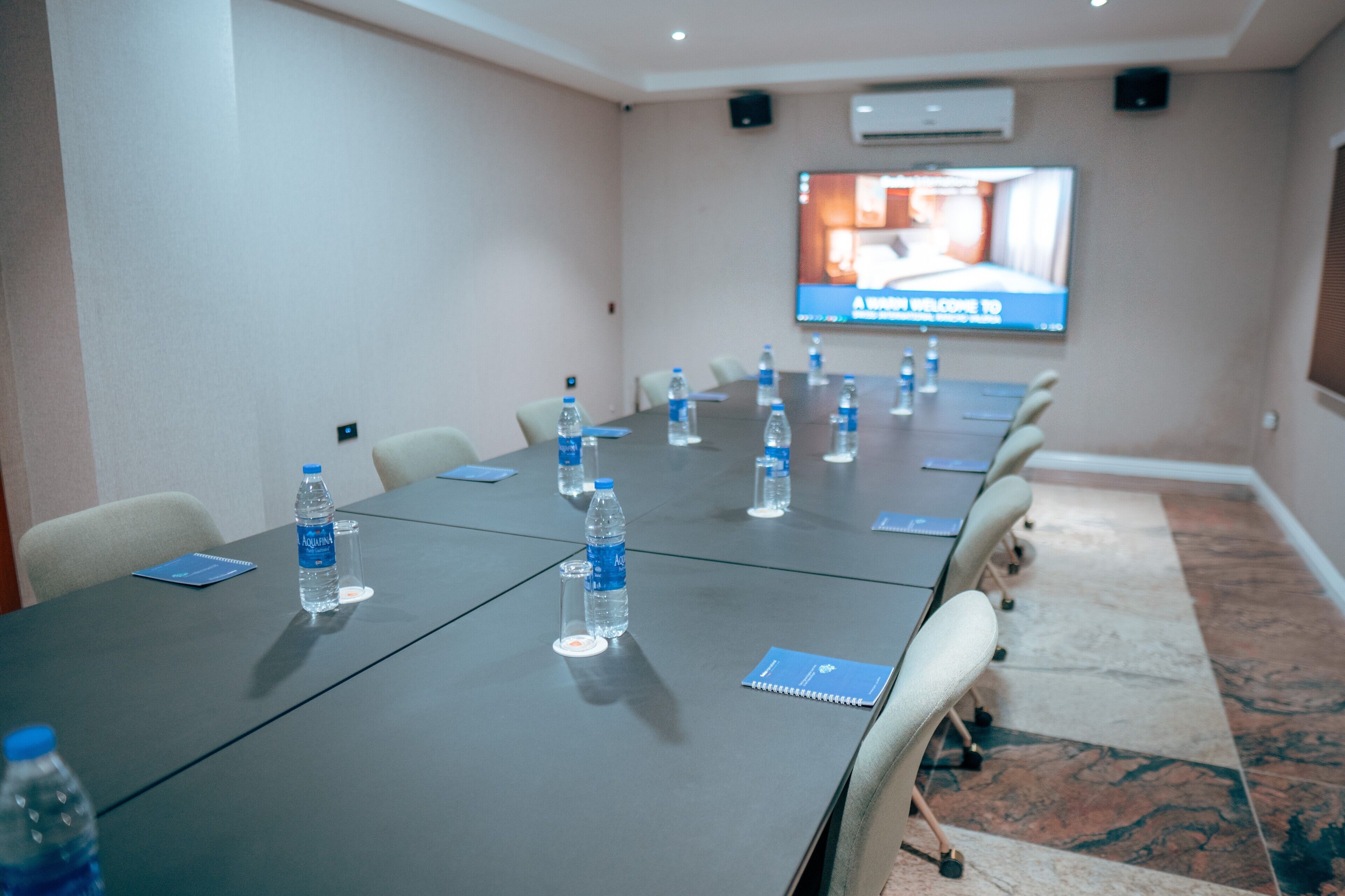 A conference room with a long table, chairs, water bottles, and a TV screen displaying a welcome message.