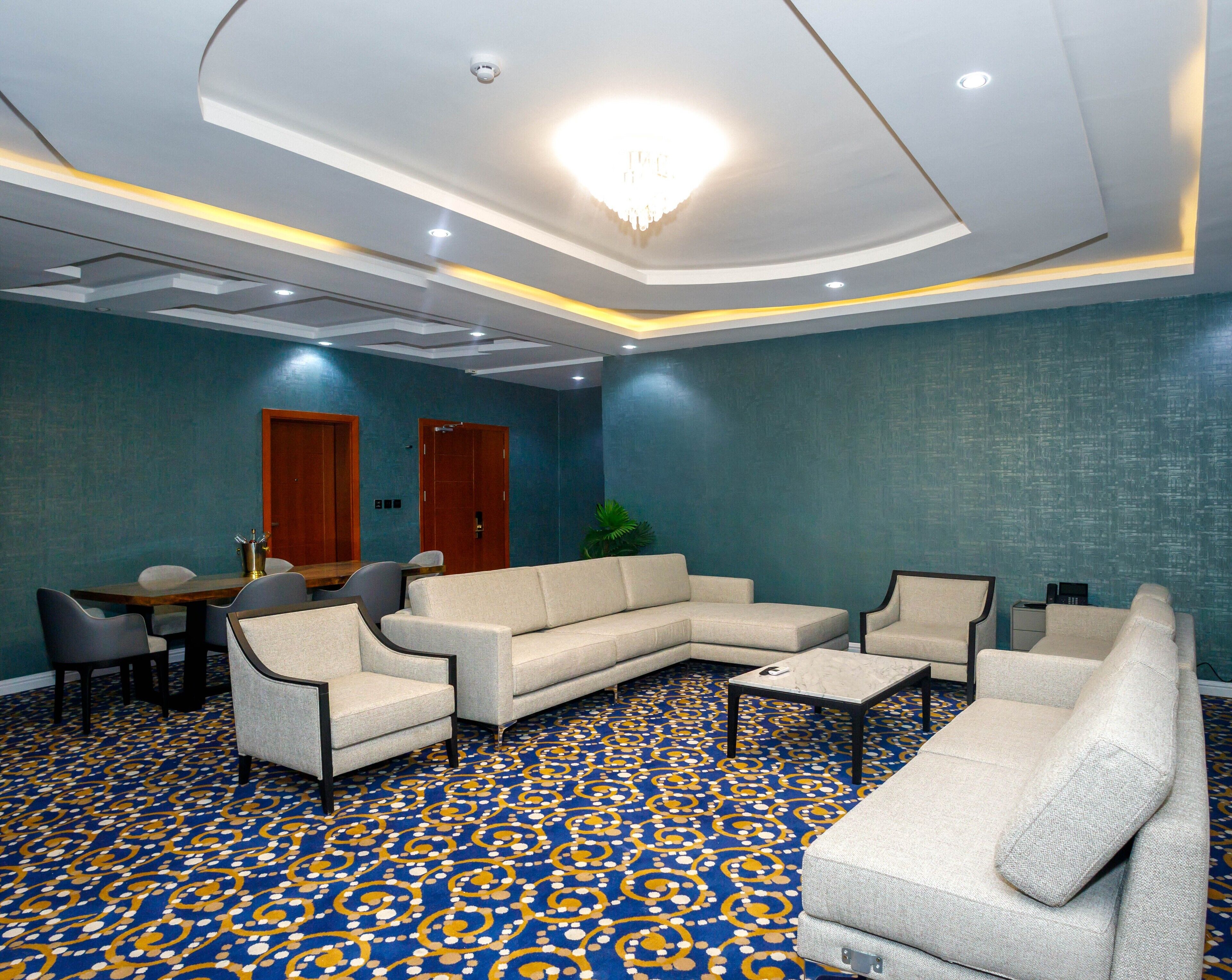 A hotel room with a large L-shaped sofa, a smaller sofa, and a coffee table. There is a dining table with chairs, a potted plant, and a chandelier.