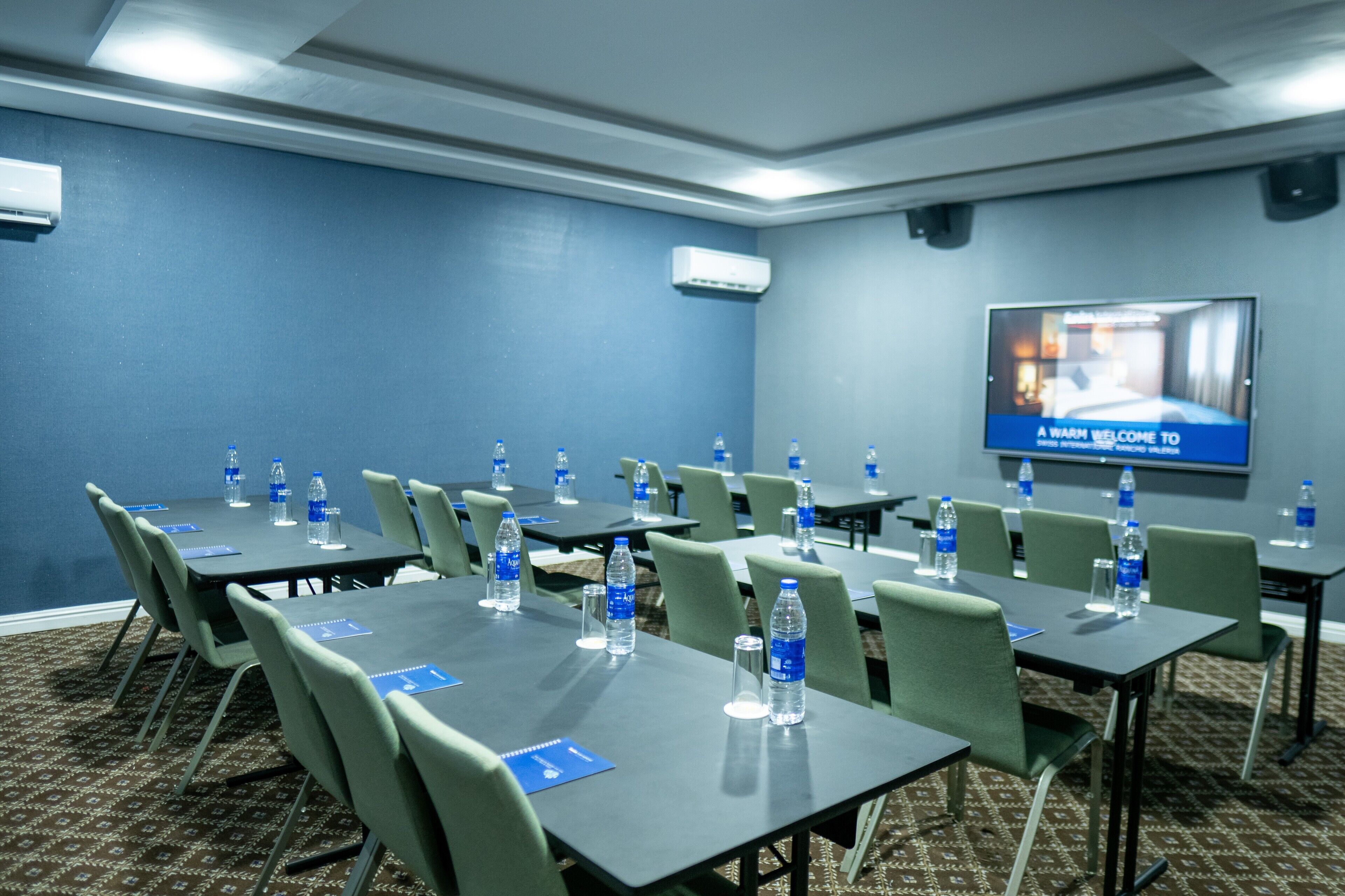 A conference room with multiple tables, chairs, water bottles, and a television screen displaying a welcome message.
