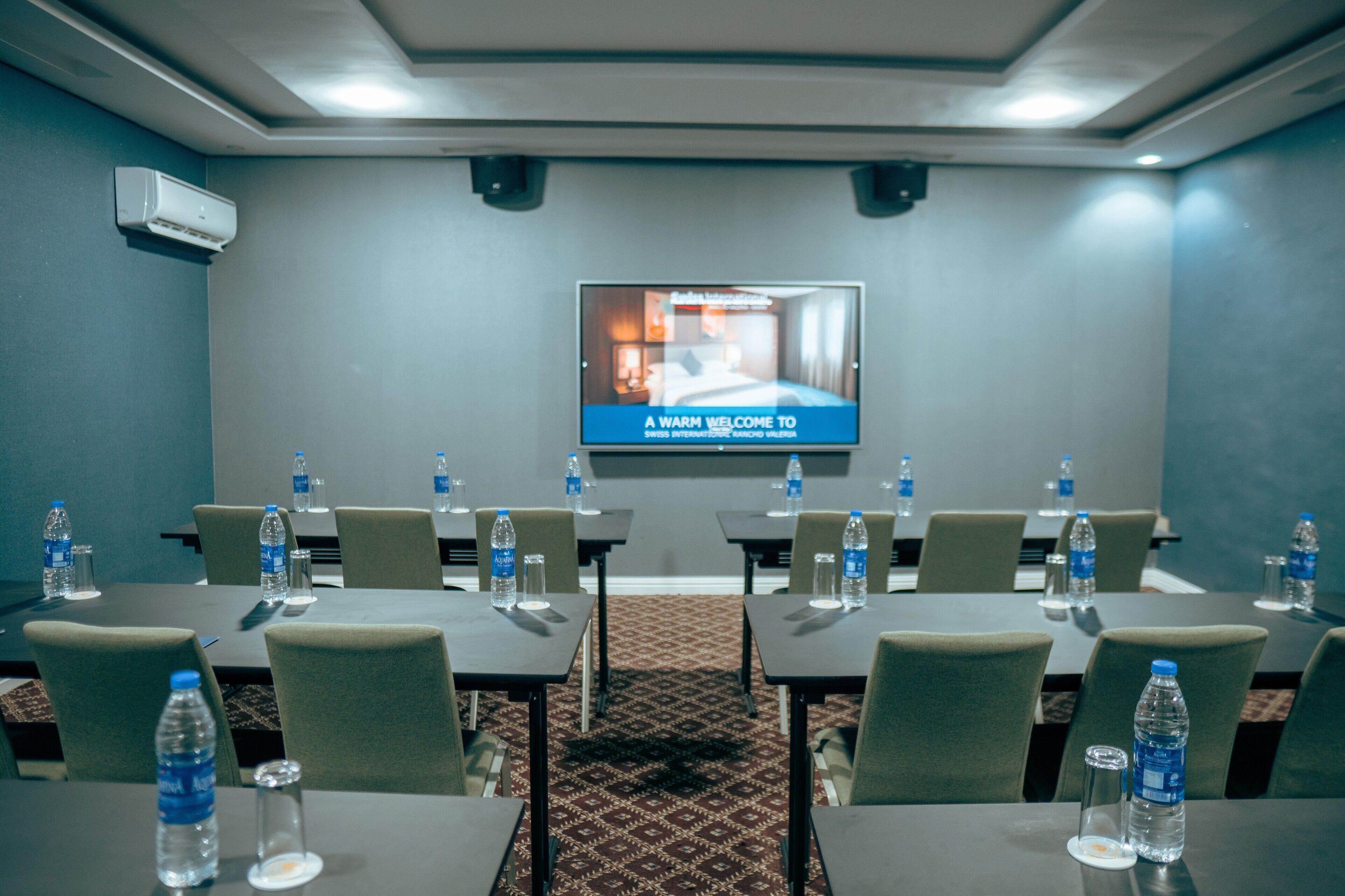 A conference room with tables, chairs, water bottles, and a television screen displaying a welcome message.