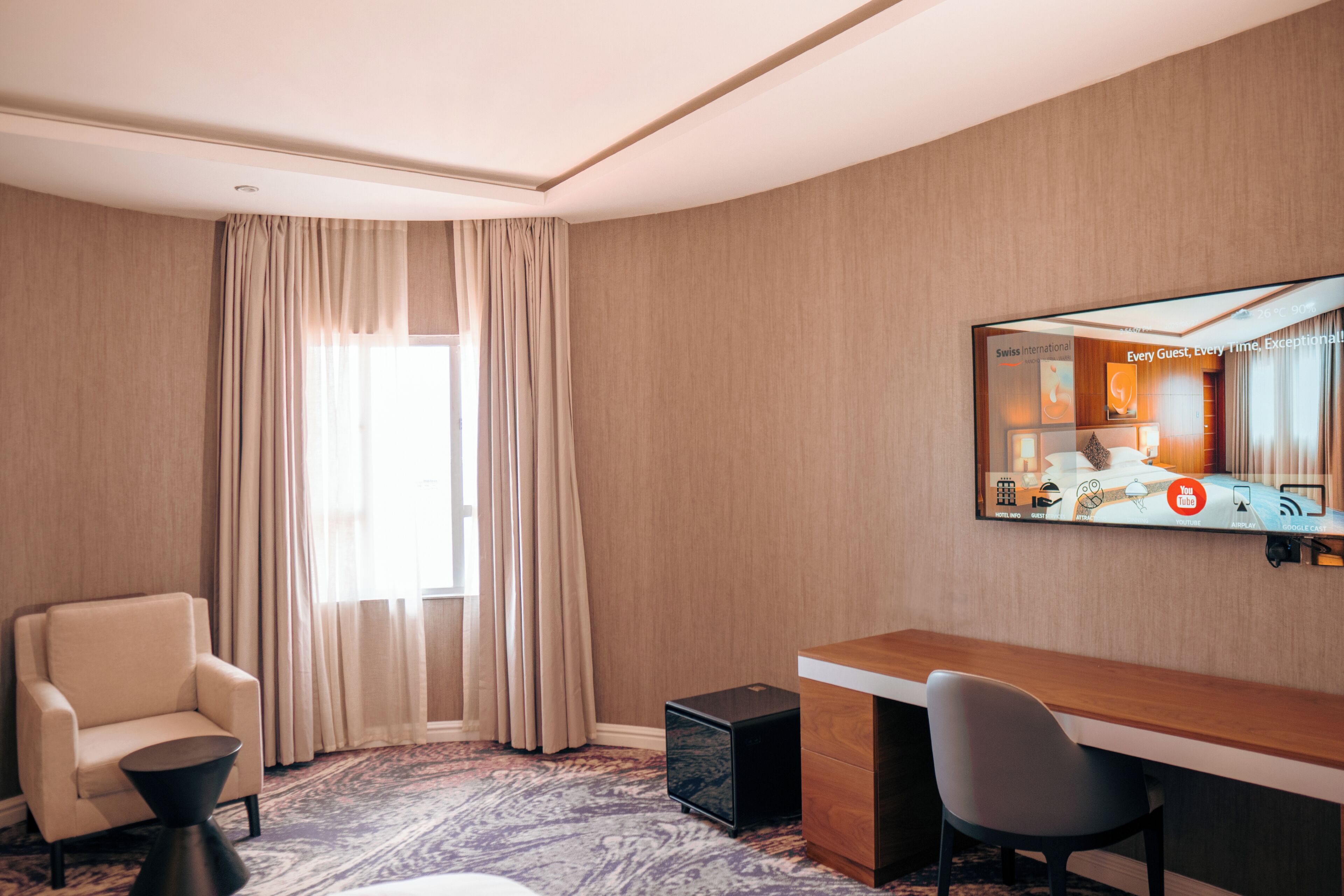 A hotel room with a large window, a desk, a chair, a small table, and a speaker.