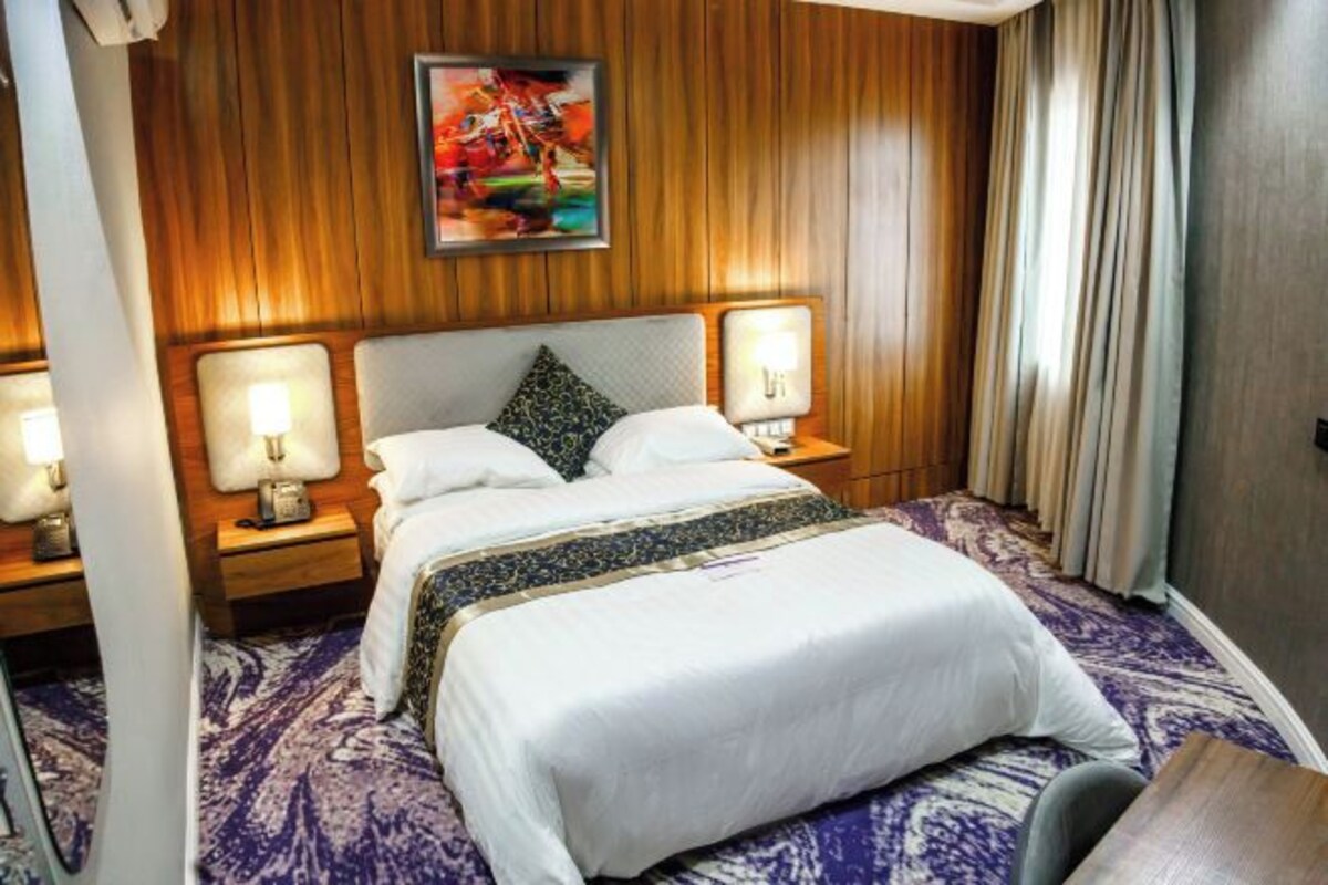 A hotel room with a large bed, two bedside tables with lamps, a headboard with a painting, and a patterned carpet.