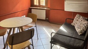 A room with a round table, two chairs, a sofa with a pillow, a window with blinds, and an air conditioning unit. - VERONA EASY LOFT - ID M0230911208 (Verona)