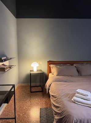 Een slaapkamer met een bed, nachtkastje met lamp, een klein plankje met boeken en een vloerkleed.