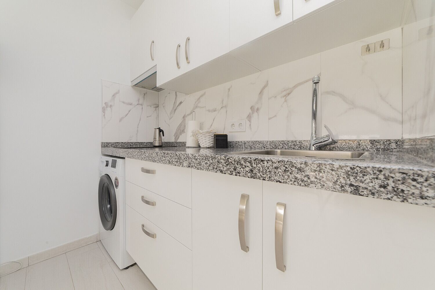 Cocina moderna con armarios blancos, encimera de granito, lavadora integrada y pileta de acero inoxidable.