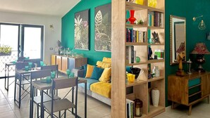 A living room with a teal wall, a wooden bookshelf, a sofa with yellow cushions, and a dining area with chairs and a table. - Rooftop53  (Catania)